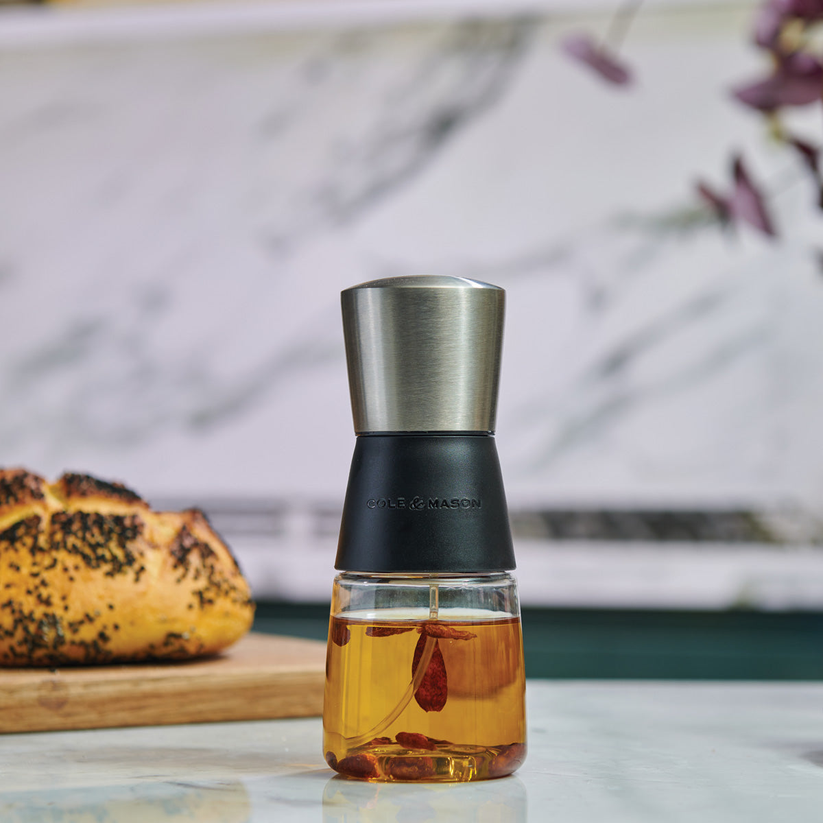 A glass and stainless steel oil infuser filled with oil and herbs sits on a counter, with a loaf of bread on a wooden board in the background. The scene has a modern kitchen setting.