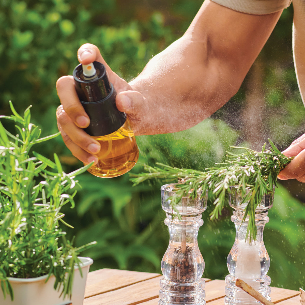 A person sprays oil from a glass sprayer onto fresh rosemary held over salt and pepper grinders on a wooden table outdoors, with green plants in the background.