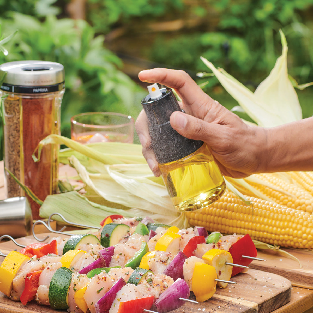 A hand sprays oil onto colorful skewers of vegetables and chicken on a wooden table outdoors. Fresh corn, spice jars, and greenery are visible in the background.