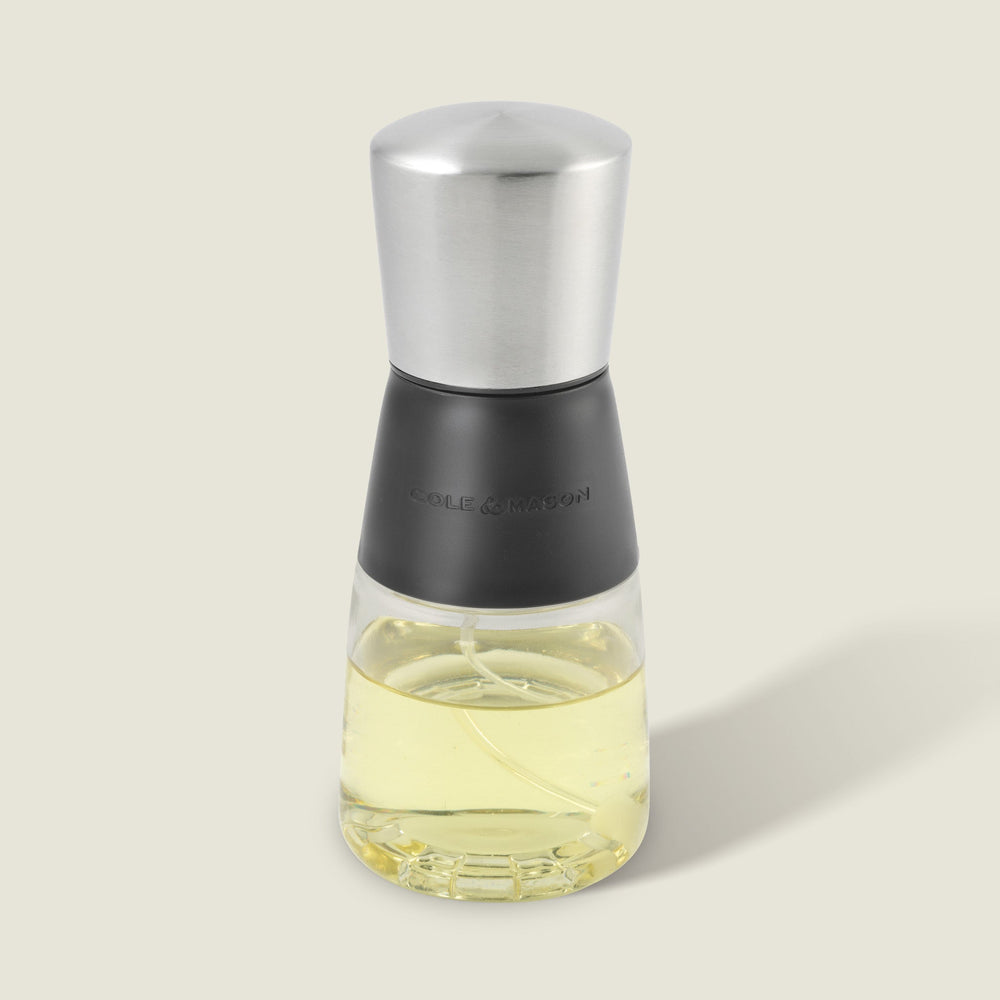 The Cole & Mason Epping Oil & Vinegar Mister features a stainless steel top, black middle section, and glass body filled with light yellow oil. Its fine misting mechanism helps promote healthier cooking, all set against a plain light background.