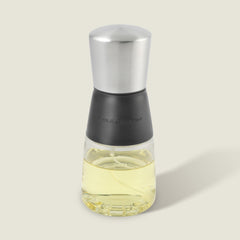 The Cole & Mason Epping Oil & Vinegar Mister features a stainless steel top, black middle section, and glass body filled with light yellow oil. Its fine misting mechanism helps promote healthier cooking, all set against a plain light background.