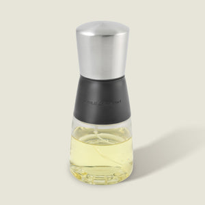 The Cole & Mason Epping Oil & Vinegar Mister features a stainless steel top, black middle section, and glass body filled with light yellow oil. Its fine misting mechanism helps promote healthier cooking, all set against a plain light background.