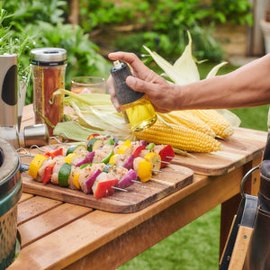 Using the Cole & Mason Epping Oil & Vinegar Mister, a person sprays a fine mist of oil over veggie skewers with bell peppers, onions, and zucchini beside fresh corn on the cob outdoors for healthier grilling.
