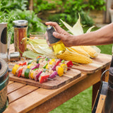 Using the Cole & Mason Epping Oil & Vinegar Mister, a person sprays a fine mist of oil over veggie skewers with bell peppers, onions, and zucchini beside fresh corn on the cob outdoors for healthier grilling.
