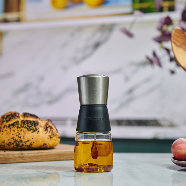 The Cole & Mason Epping Oil & Vinegar Mister with a stainless steel top sits on the counter, encouraging healthier meals, while a loaf of bread on a wooden board and fruit rest nearby against a marble backsplash.