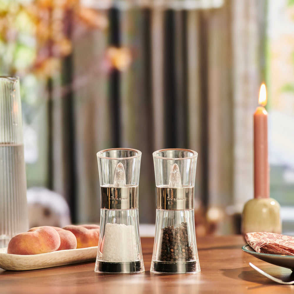 The Cole & Mason Flip Salt & Pepper Mill Set features two transparent mills with silver bands, one filled with Belgian sea salt and the other with Vietnamese peppercorns, displayed side by side on a light background.