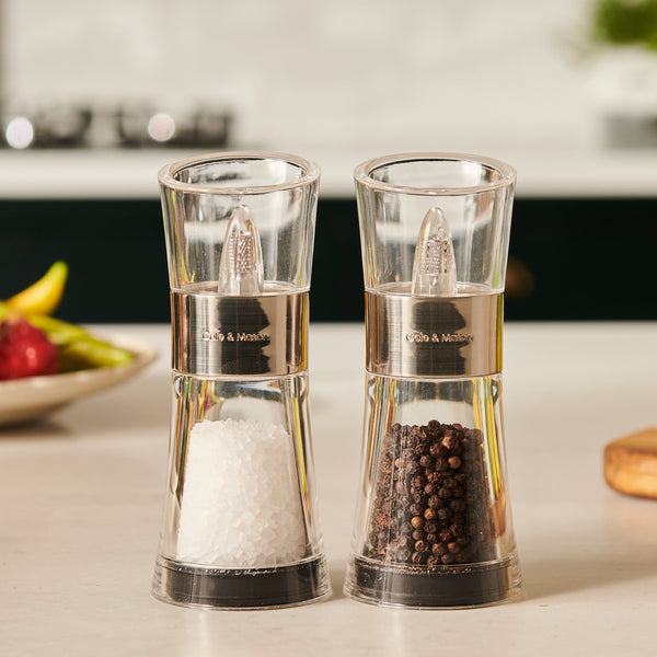 The Cole & Mason Flip Salt & Pepper Mill Set features two transparent mills, one with gourmet Belgian sea salt and the other with whole Vietnamese peppercorns, displayed side by side on a kitchen counter with blurred kitchen items in the background.