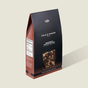 A box of Cole & Mason Gourmet Pepper Box features a clear window displaying mixed peppercorns. The mostly black packaging with brown accents is perfect for grinding fresh flavor and includes a nutrition facts label on the side.