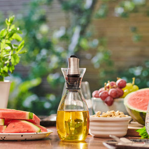 The Cole & Mason Haverhill Oil & Vinegar Pourer with precision spout rests on an outdoor table among watermelon slices, nuts, grapes, and fresh herbs, with a leafy garden softly blurred in the background.
