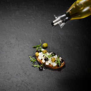 A slice of bread with feta, olives, and arugula is drizzled with olive oil from the Cole & Mason Haverhill Oil & Vinegar Pourer with a precision spout, shown on a dark stone surface.
