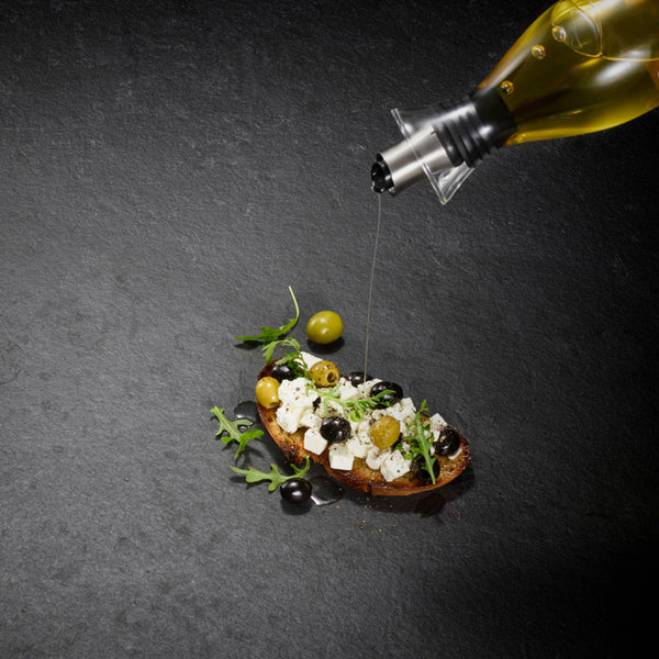A slice of bread with feta, olives, and arugula is drizzled with olive oil from the Cole & Mason Haverhill Oil & Vinegar Pourer with a precision spout, shown on a dark stone surface.