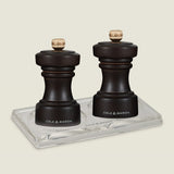 The Cole & Mason Hoxton Chocolate Wood Mill & Acrylic Tray Set adds subtle Art Deco charm to any table setting, featuring two dark wooden mills with gold knobs elegantly displayed on a clear acrylic tray.