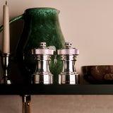 The Cole & Mason Hoxton Silver Plated Salt & Pepper Mill Set gleams on a dark shelf, accompanied by a tall white candle, green ceramic pitcher, and wooden bowl—stylish essentials for elegant dining.