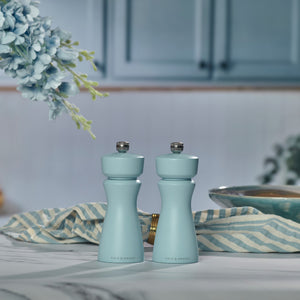 Two refillable Kenton Blue Salt & Pepper Mills by Cole & Mason sit on a marble countertop with a striped cloth, bowl, and blue cabinets in the background. Flowers hang from the upper left corner.