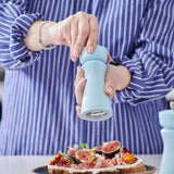 Wearing a blue and white striped shirt, a person seasons food topped with figs and edible flowers using the Cole & Mason Kenton Salt & Pepper Mill in Blue Salt, featuring a durable ceramic mechanism.