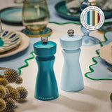 The Cole & Mason Kenton Salt & Pepper Mills in Blue Salt, featuring a durable ceramic mechanism, are displayed on a set dining table with plates, cutlery, plants, and a color selection graphic in the top right corner.