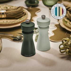Two elegant Cole & Mason Kenton Salt & Pepper Mills in Rosemary, one dark and one light green, sit on a stylish dining table with plates and cutlery. A badge highlights their adaptable design and mentions six available color options.