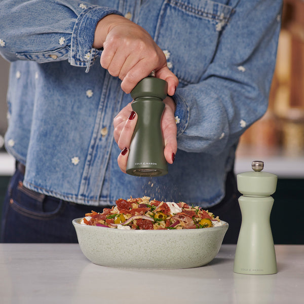 A person in a denim shirt grinds pepper over salad with a Cole & Mason Kenton Rosemary Salt & Pepper Mill featuring a ceramic mechanism, while another matching mill with interchangeable knobs sits on the countertop.