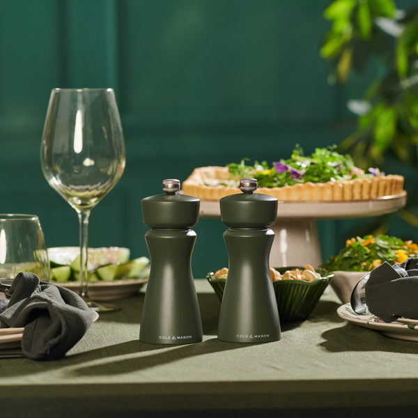 Two Cole & Mason Kenton Salt & Pepper Mills in Rosemary with ceramic mechanisms are on a dining table set with wine glasses, plates, napkins, and a vegetable tart, accented by greenery against a dark green backdrop.