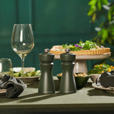 Two Cole & Mason Kenton Salt & Pepper Mills in Rosemary with ceramic mechanisms are on a dining table set with wine glasses, plates, napkins, and a vegetable tart, accented by greenery against a dark green backdrop.
