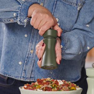 A person in a denim shirt grinds a Cole & Mason Kenton Salt & Pepper Mill in Rosemary with a ceramic mechanism over a bowl of salad, adding freshly ground pepper.