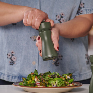 A person in a blue floral shirt grinds pepper over a salad of leafy greens, nuts, and vegetables using the Cole & Mason Kenton Salt & Pepper Mill in Rosemary, which features a durable ceramic mechanism.