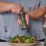 A person in a blue floral shirt grinds pepper over a salad of leafy greens, nuts, and vegetables using the Cole & Mason Kenton Salt & Pepper Mill in Rosemary, which features a durable ceramic mechanism.
