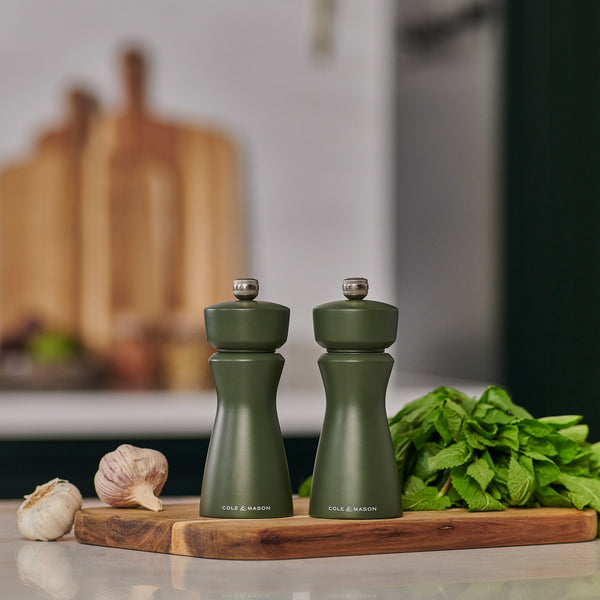 Two Cole & Mason Kenton Salt & Pepper Mills in Rosemary, featuring durable ceramic mechanisms, sit on a wooden board beside fresh mint and garlic, bringing adaptable style to a modern kitchen with blurred cutting boards behind.