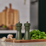 Two Cole & Mason Kenton Salt & Pepper Mills in Rosemary, featuring durable ceramic mechanisms, sit on a wooden board beside fresh mint and garlic, bringing adaptable style to a modern kitchen with blurred cutting boards behind.