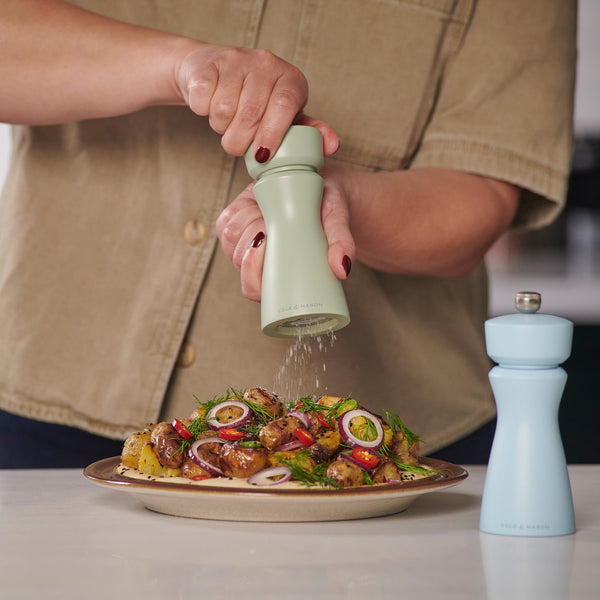 A person seasons roasted vegetables with the pale green Cole & Mason Kenton Sage Salt & Pepper Mill, featuring a ceramic mechanism and interchangeable knobs. A light blue mill sits nearby as they enjoy both style and function in a tan shirt.