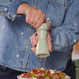 A person in a denim shirt grinds seasoning from a Cole & Mason Kenton Salt & Pepper Mill in Sage with interchangeable knobs over a colorful salad bowl.