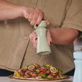 A person seasons herb-roasted potatoes and onions with the Cole & Mason Kenton Salt & Pepper Mill in Sage, which features a ceramic mechanism. Next to it is a light blue grinder with interchangeable knobs. The person is wearing a beige shirt.