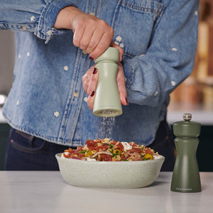 Wearing a denim jacket, a person grinds pepper onto salad with a Cole & Mason Kenton Sage Salt & Pepper Mill, featuring a ceramic mechanism, as another green grinder with interchangeable knobs rests nearby on the white countertop.