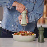 Wearing a denim jacket, a person grinds pepper onto salad with a Cole & Mason Kenton Sage Salt & Pepper Mill, featuring a ceramic mechanism, as another green grinder with interchangeable knobs rests nearby on the white countertop.