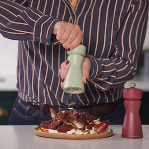A person in a striped shirt grinds pepper onto food with the Cole & Mason Kenton Sage Salt & Pepper Mill, featuring a ceramic mechanism. A red grinder with interchangeable knobs rests on the white countertop nearby.