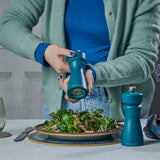A person in a teal sweater grinds pepper onto salad with the Cole & Mason Kenton Sea Salt Blue Salt & Pepper Mill, while the matching set sits on the table—a stylish addition to contemporary kitchens.