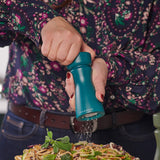 A person in a floral shirt seasons their food using the Cole & Mason Kenton Salt & Pepper Mill in Sea Salt, featuring a ceramic mechanism—pepper falls onto the dish, making it a stylish pick for contemporary kitchens.