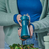 A person with teal nails uses the Cole & Mason Kenton Salt & Pepper Mill in Sea Salt to season a salad. The mill features a ceramic mechanism, perfect for sea salt in modern kitchens. Theyre wearing a blue shirt and light green cardigan.