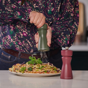 Wearing a floral shirt, a person grinds black pepper onto pasta with a green Cole & Mason Kenton Rosemary Salt & Pepper Mill featuring a ceramic mechanism, while a red grinder with interchangeable knobs sits on the table.