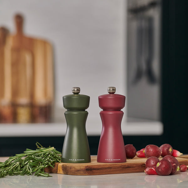 Two Cole & Mason Kenton Rosemary Salt & Pepper Mills with interchangeable knobs and a ceramic mechanism sit on a wooden board with rosemary and radishes in a modern kitchen, blurred cutting boards in the background.
