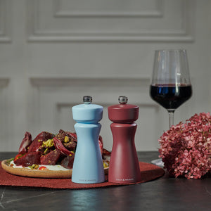 Two Cole & Mason Kenton Sichuan Pepper Red Salt & Pepper Mills, one light blue and one maroon, are on a table with Sichuan pepper, a plate of food, red wine, pink flowers, and a white decorative wall in the background.