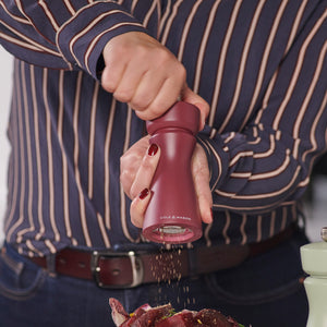A person in a striped shirt grinds pepper onto food with a red Cole & Mason Kenton Salt & Pepper Mill in Sichuan Pepper, featuring a ceramic mechanism. Only their torso and hands are visible.
