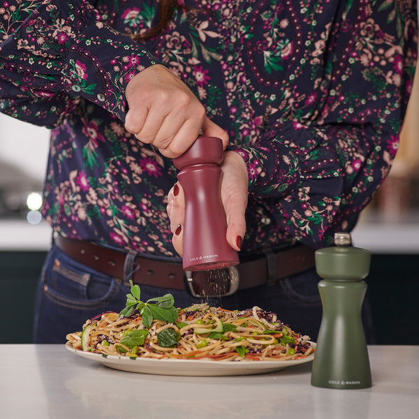 A person in a floral shirt grinds pepper onto pasta using the Cole & Mason Kenton Sichuan Pepper Red Salt & Pepper Mill with a ceramic mechanism, while another green grinder rests on the table nearby.