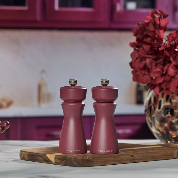 Two matching Cole & Mason Kenton Sichuan Pepper Red Salt & Pepper Mills with durable ceramic mechanisms sit on a wooden tray on a marble countertop, framed by red cabinets and a vase of burgundy flowers in the background.