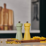 The Cole & Mason Kenton Salt & Pepper Mill in Turmeric Yellow, with a ceramic mechanism, sits on a wooden board beside sliced yellow squash and pistachios. A green mill and blurred cutting boards are in the background.