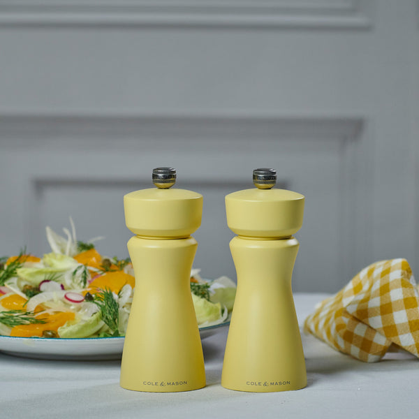 Two Cole & Mason Kenton Salt & Pepper Mills in turmeric yellow, featuring a ceramic mechanism, sit on a table with a plate of salad and a yellow checkered napkin in the background.