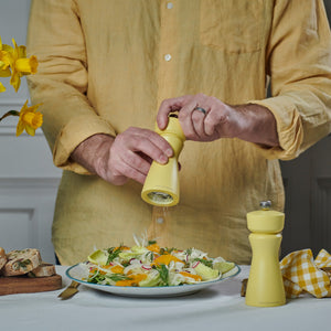 Wearing a yellow shirt, someone grinds pepper onto a fresh salad with the Cole & Mason Kenton Salt & Pepper Mill in Turmeric Yellow at a table set with bread and yellow flowers, creating a cheerful spring-themed atmosphere.