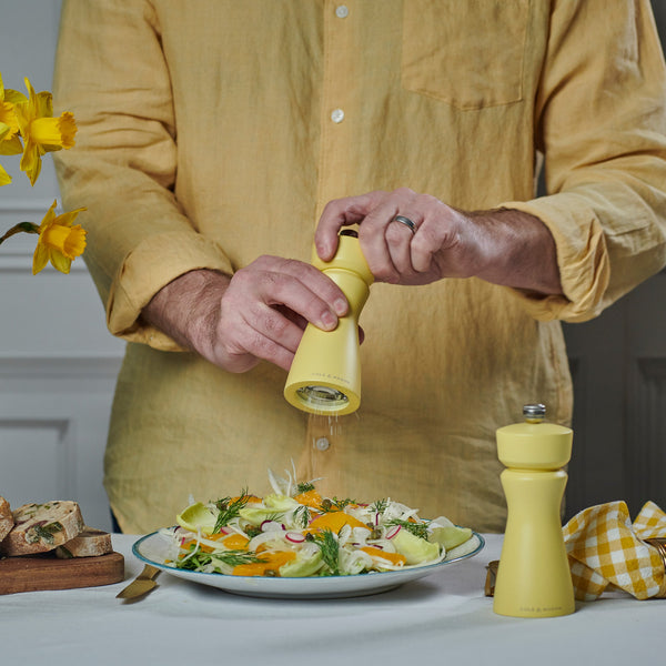 Wearing a yellow shirt, someone grinds pepper onto a fresh salad with the Cole & Mason Kenton Salt & Pepper Mill in Turmeric Yellow at a table set with bread and yellow flowers, creating a cheerful spring-themed atmosphere.