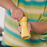 A person in a striped sweater uses the Cole & Mason Kenton Salt & Pepper Mill in Turmeric, featuring a durable ceramic mechanism, as freshly ground pepper falls from the bottom.