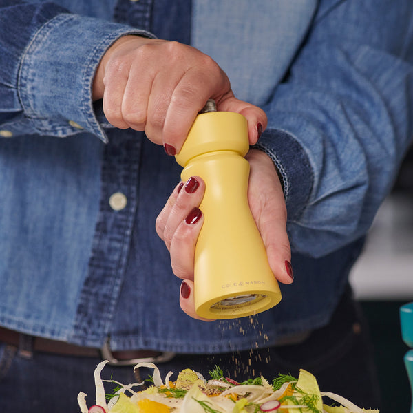 A person in a denim shirt with red-painted nails seasons a vegetable-topped dish using the Cole & Mason Kenton Salt & Pepper Mill in Turmeric, featuring a ceramic mechanism.